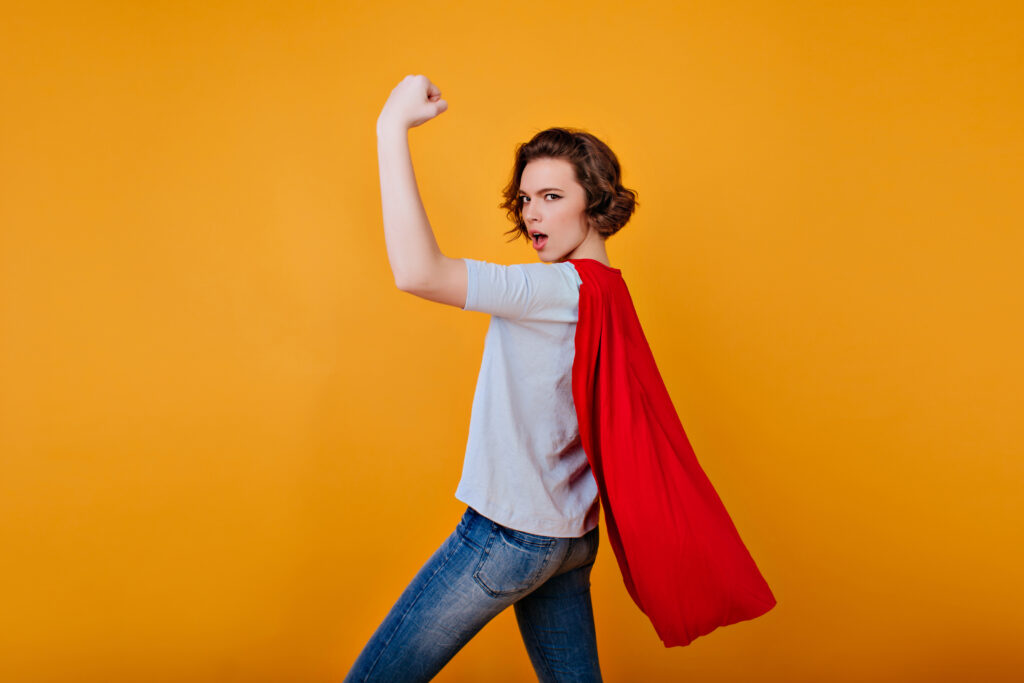 Strong girl in denim pants and blue t-shirt posing with hand up. Funny female model in red cloak showing her muscles on bright background..